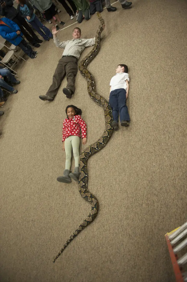 Two children laying on the floor next to a snake.