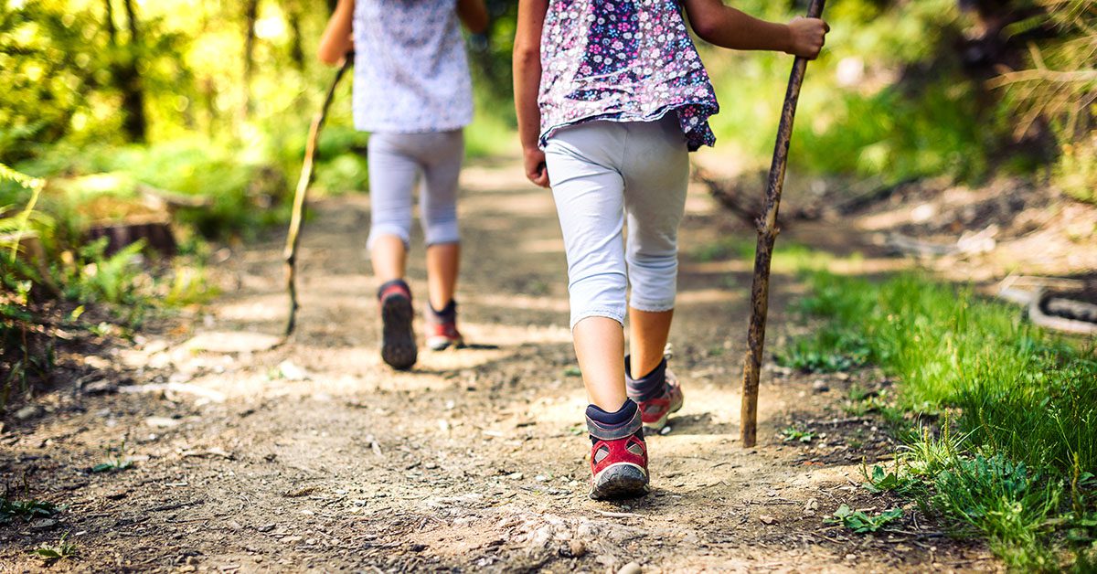 Children hiking in mountains or forest with sport hiking shoes. Girls are walking trough forest path wearing mountain boots and walking sticks. Frog perspective with focus on the shoes.